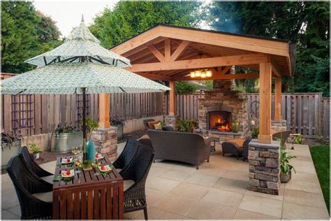 Timber-frame covered pavilion with a stone fireplace, stone-clad columns, and a dining table set on a paver patio at dusk