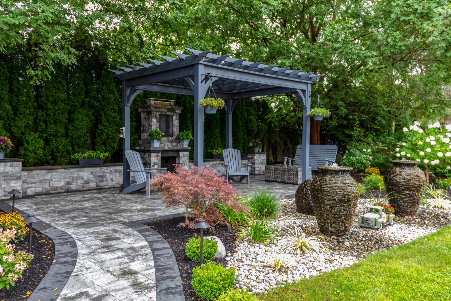 Backyard outdoor living space with a pergola, stone fireplace, paver patio, and layered plantings